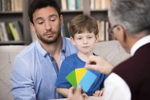 Little boy and his father talking with psychologist. Psychologist making test with boy. There are many books in psychologist office